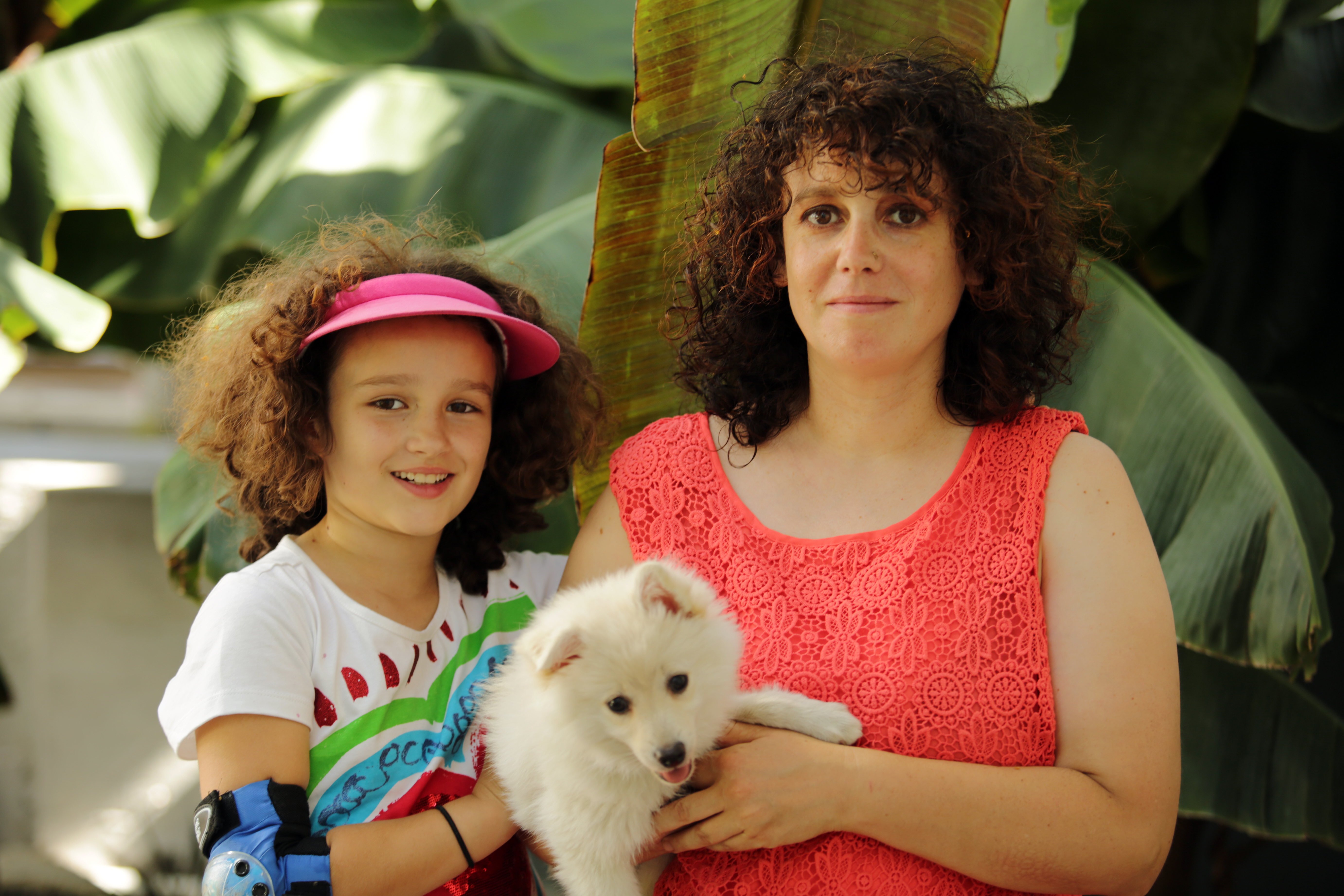 Mädchen mit braunen Locken und buntem T-Shirt neben Frau mit braunen Locken und rotem Top einen Hund auf dem Arm haltend