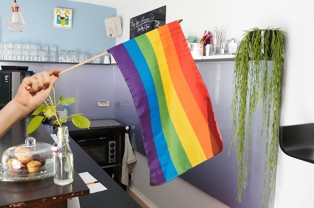 Hand hält von links eine Regenbogenfahne ins Bild, im Hintergrund steht die Bar eines Cafés.