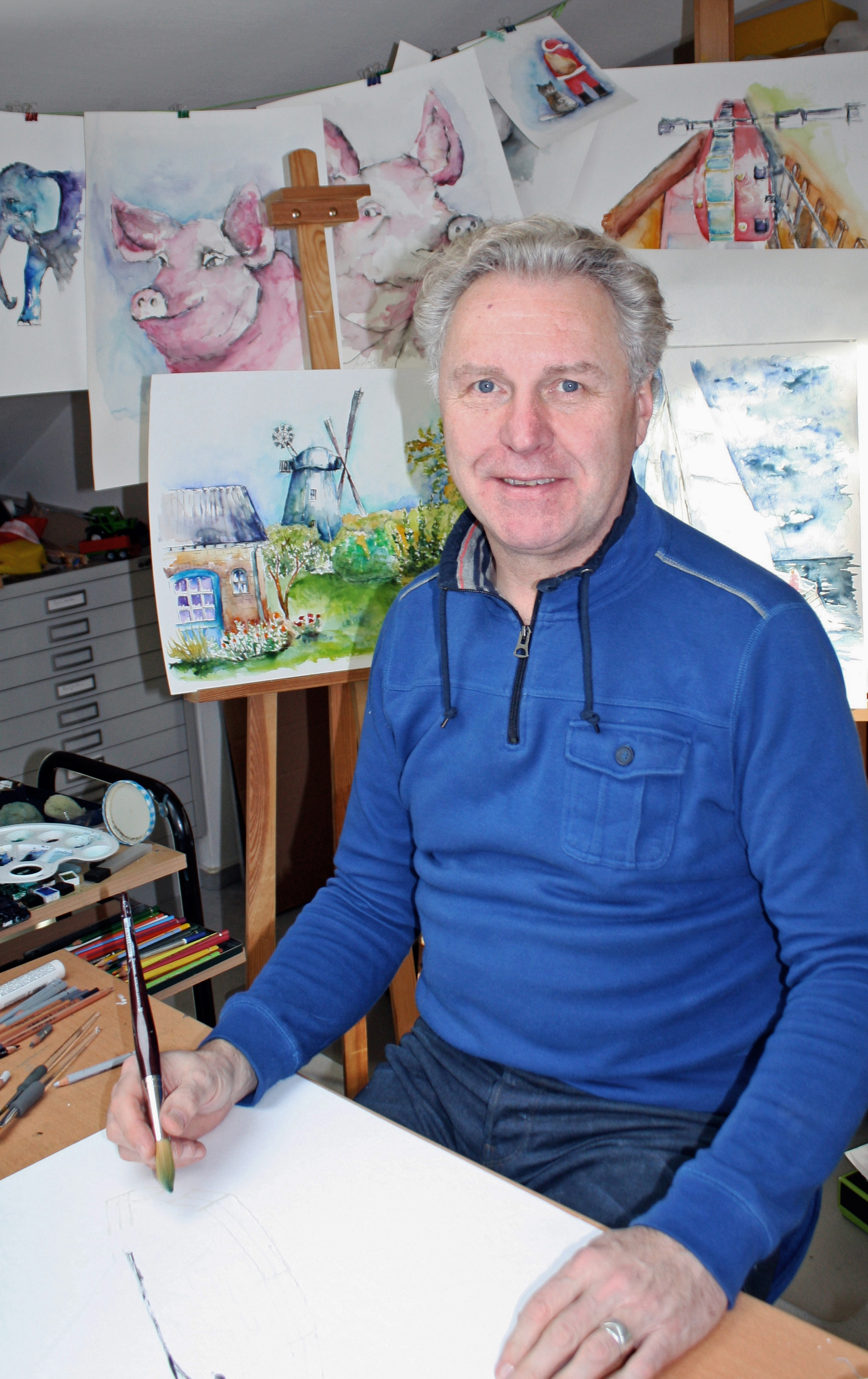 Mann sitzt mit Pinsel vor einem leeren Blatt. Im Hintergrund hängen Aquarellbilder mit unterschiedlichen Motiven.