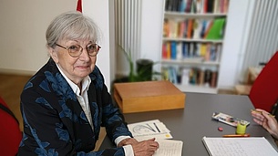 Frau mit grauen Haaren, Brille und blauem Blazer sitzt an einem Tisch, auf dem ein Kasten mit Buchstabenplatten steht