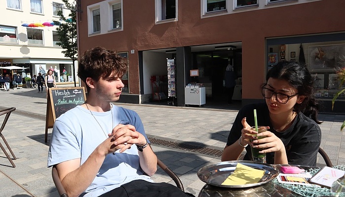 Eine junge Frau und ein junger Mann sitzen an einem Bistrotisch und trinken Smoothies.