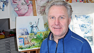 Mann sitzt mit Pinsel vor einem leeren Blatt. Im Hintergrund hängen Aquarellbilder mit unterschiedlichen Motiven.