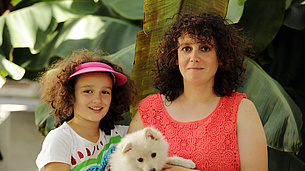 Mädchen mit braunen Locken und buntem T-Shirt neben Frau mit braunen Locken und rotem Top einen Hund auf dem Arm haltend