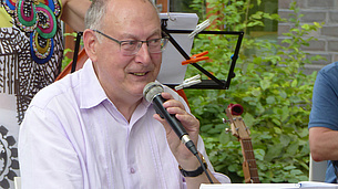 Ein älterer Mann sitzt draußen auf einem Stuhl und singt in ein Mirkofon. Vor ihm steht ein Notenständer. 