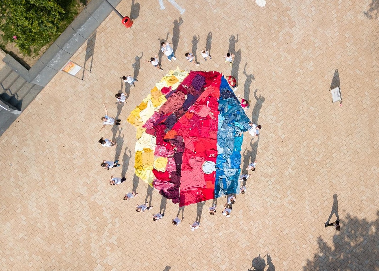 Aufnahme von oben: Viele Menschen in weißen T-Shirts stehen um ein aus gefärbtem Stoff auf den Boden gelegtes Mehrgenerationenhaus-Logo
